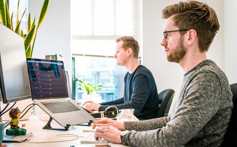 Two men working on computers in a bright office with large windows.